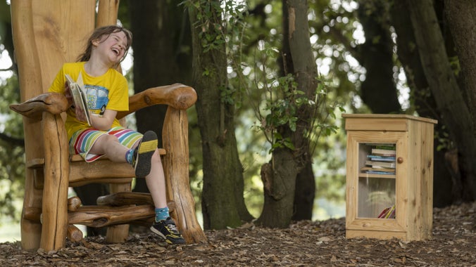 A young girl smiling and laughing as she reads under the storytelling tree at Sutton Hoo